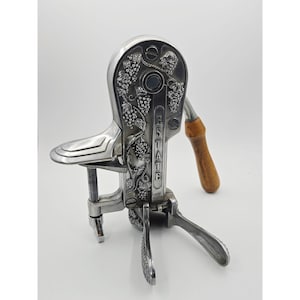 May include: A silver-toned metal apple peeler with a clamp base, featuring an embossed grape vine pattern on the main body and the word ESTATE vertically printed on the side. It includes a wooden handle and a threaded metal screw mechanism.