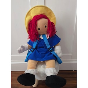 May include: A plush doll with bright red yarn hair, wearing a yellow hat, a blue dress with a white collar, and blue straps. The doll has white gloves, white socks with lace trim, and black shoes. The doll features a tan face with simple black eyes.