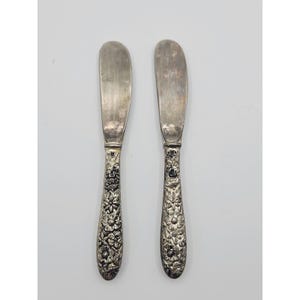 May include: Two silver-toned butter knives featuring smooth, rounded blades and handles intricately embossed with a detailed floral pattern. The metal surfaces exhibit a polished finish with darkened accents within the recessed floral design.