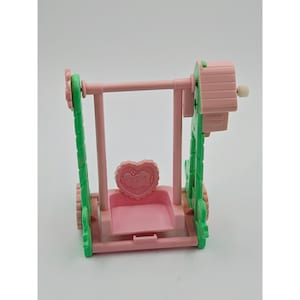May include: A toy swing set featuring a pink seat with a heart-shaped backrest, suspended by a pink horizontal bar between two green vertical support frames. The structure includes a small pink roof-like attachment on the right side.
