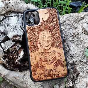 May include: A black phone case with a wood-grain design featuring a character from the horror film "Hellraiser". The character is depicted with a large, intricate headpiece and a menacing expression. The case has a cutout for the phone's camera lens.