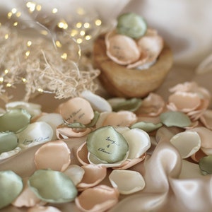 Puede incluir: Esparcidos sobre una superficie de tela beige, hay pétalos de flores de seda de color verde, melocotón y blanco. Algunos de los pétalos tienen los nombres "Joey & Chance" escritos en tinta negra.