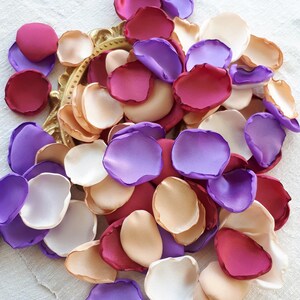 Pode incluir: Um monte de pétalas de flores de seda em tons de roxo, bordô e pêssego. As pétalas estão espalhadas sobre uma superfície de linho branco.