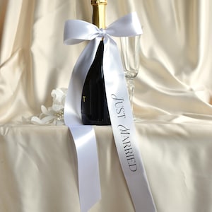 May include: A dark glass bottle with a gold foil neck wrap, adorned with a white satin ribbon tied in a bow. The ribbon features the black printed text "JUST MARRIED" flowing down the front. A clear glass flute stands beside the bottle.