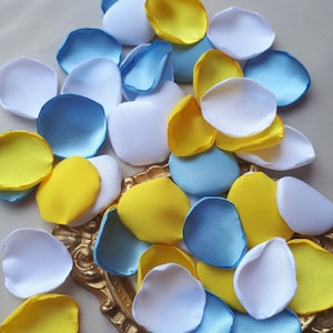 Può includere: Una collezione di petali di fiori in tessuto blu, giallo e bianco. I petali sono disposti in un mucchio su una superficie dorata.