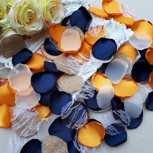 Può includere: Una collezione di petali di tessuto colorati in blu navy, arancione, pesca e bianco, con accenti di juta e pizzo. I petali sono sparsi, creando un'esposizione strutturata e decorativa, adatta per matrimoni o lavori artigianali.