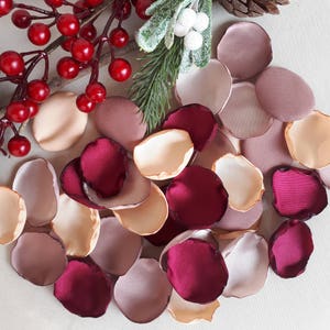 Può includere: Assortimento di petali di rosa in raso color borgogna, rosa cipria e crema. I petali sono sparsi su una superficie bianca.