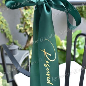 May include: A dark green satin ribbon tied in a bow around a black chair back. The ribbon features the word Reserved printed in a metallic gold script font vertically along the center of the fabric, creating a decorative sign for seating.