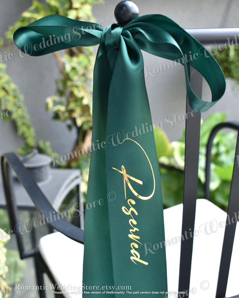 May include: A dark green satin ribbon tied in a bow around a black chair back. The ribbon features the word Reserved printed in a metallic gold script font vertically along the center of the fabric, creating a decorative sign for seating.
