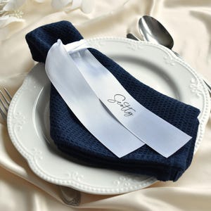 May include: A navy blue waffle-textured napkin folded on a white scalloped-edge plate, secured with a white satin ribbon featuring the name Scotty printed in black script. A silver fork and spoon are placed beside the plate on a cream fabric.