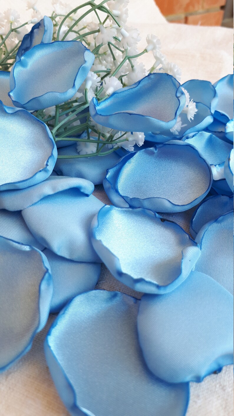 May include: A scattering of light blue satin rose petals. The petals are arranged in a random pattern on a white surface.
