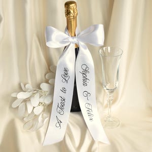 May include: A champagne bottle with a gold foil top, adorned with a white satin ribbon tied in a bow. The ribbon features black printed text reading A Toast to Love on one side and Sophia & Felix on the other, accompanied by a clear glass flute.