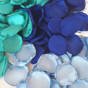 May include: A pile of teal, blue, and light blue silk rose petals. The petals are arranged in a semi-circle, with the teal petals at the top, the blue petals in the middle, and the light blue petals at the bottom.