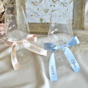 Puede incluir: Dos copas de vino transparentes con cintas de satén. Una copa tiene una cinta melocotón con "Anna" y "Bridesmaid" impresos. La otra copa tiene una cinta azul claro con "Anna" y "Bride" impresos. Un estampado floral enmarcado está en el fondo.
