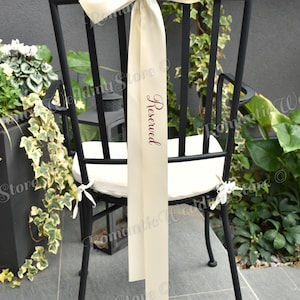 May include: A black metal chair with a white seat cushion features a long, cream-colored satin ribbon tied in a bow at the top. The ribbon hangs down the center of the backrest and displays the word Reserved printed in a dark script font.