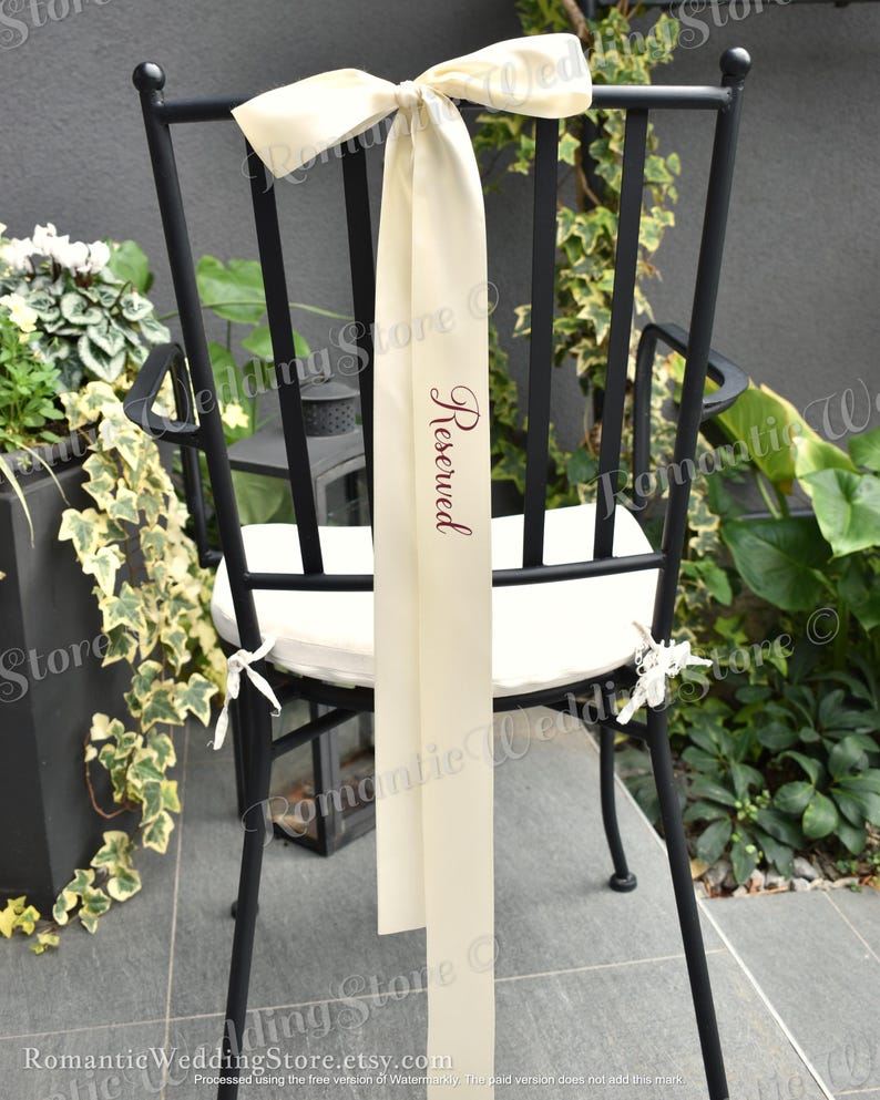 May include: A black metal chair with a white seat cushion features a long, cream-colored satin ribbon tied in a bow at the top. The ribbon hangs down the center of the backrest and displays the word Reserved printed in a dark script font.