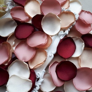 May include: A collection of silk rose petals in shades of burgundy, blush pink, ivory, and peach. The petals are arranged on a white surface with a decorative frame.