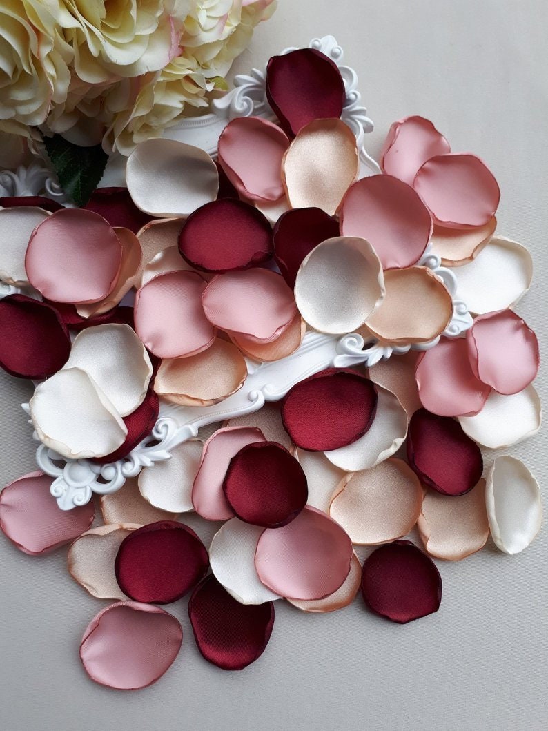 May include: Assortment of silk rose petals in ivory, blush pink, peach, and burgundy colors. The petals are scattered on a white surface.