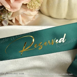 May include: A dark green satin ribbon featuring the word Reserved written in a metallic gold cursive script font. The ribbon is draped over a smooth, light-colored fabric surface, creating soft folds and a reflective sheen on the material.