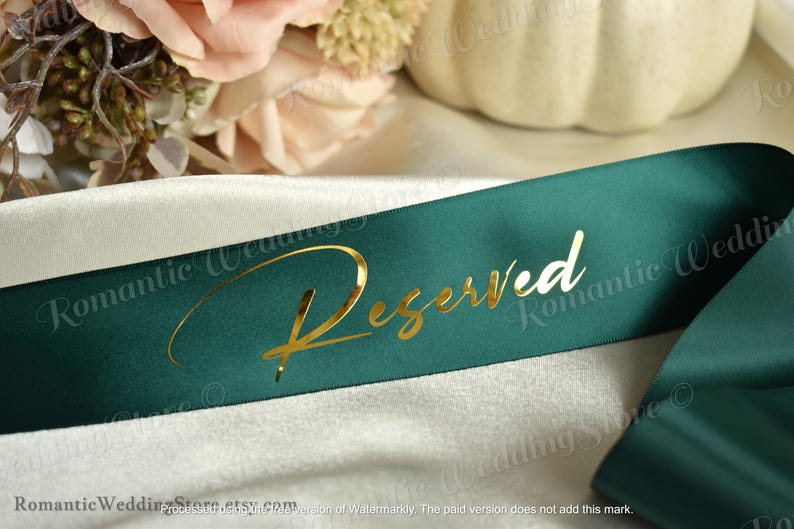 May include: A dark green satin ribbon featuring the word Reserved written in a metallic gold cursive script font. The ribbon is draped over a smooth, light-colored fabric surface, creating soft folds and a reflective sheen on the material.