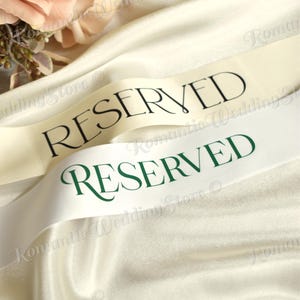 May include: Two satin ribbons draped over fabric, one cream-colored with black text reading RESERVED and one white ribbon with dark green text reading RESERVED. The ribbons have a smooth, reflective finish and are positioned near soft floral accents.
