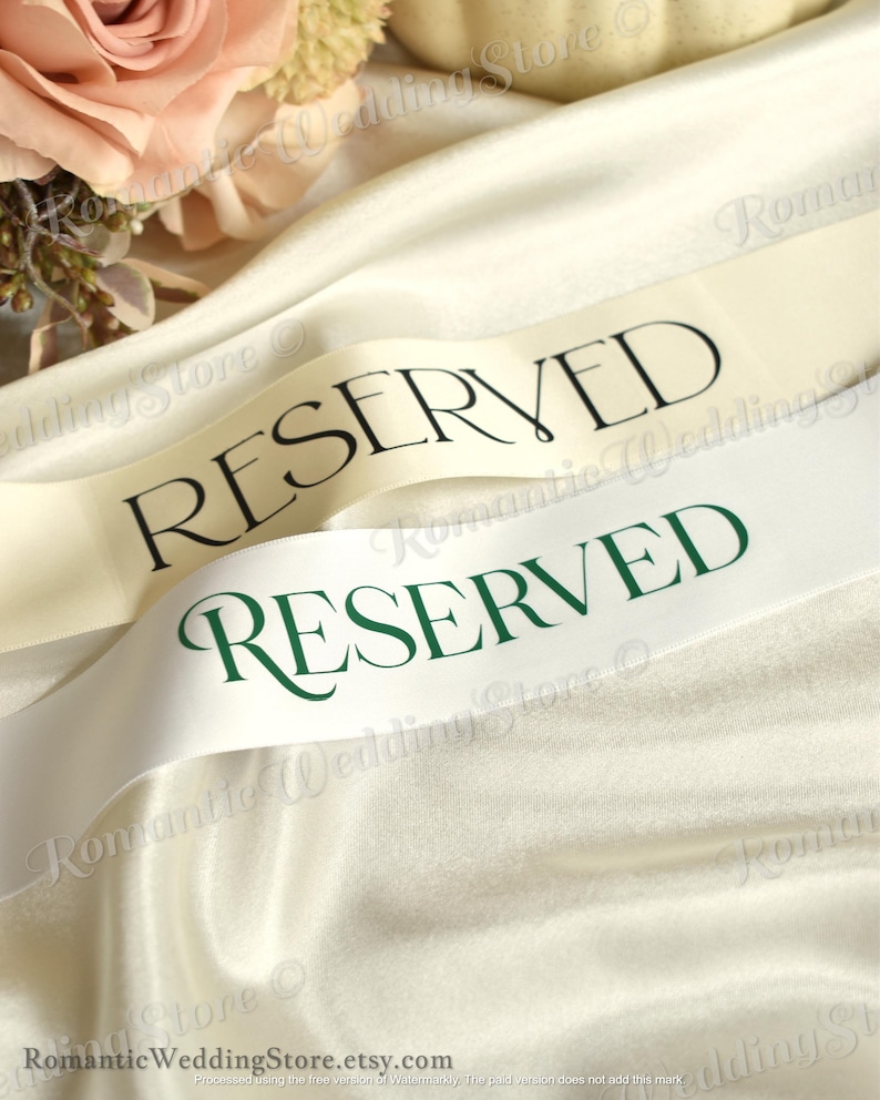 May include: Two satin ribbons draped over fabric, one cream-colored with black text reading RESERVED and one white ribbon with dark green text reading RESERVED. The ribbons have a smooth, reflective finish and are positioned near soft floral accents.