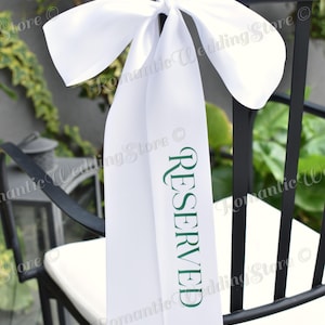May include: A white satin ribbon tied in a bow around the top of a black chair. The ribbon features the word RESERVED printed vertically in dark green capital letters. The fabric has a smooth, reflective finish and hangs down the back of the chair.