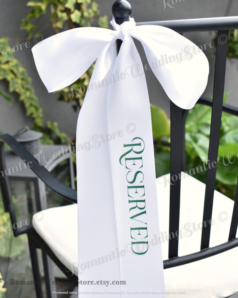 May include: A white satin ribbon tied in a bow around the top of a black chair. The ribbon features the word RESERVED printed vertically in dark green capital letters. The fabric has a smooth, reflective finish and hangs down the back of the chair.
