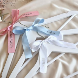 May include: Three white wooden hangers with silver hooks, each adorned with a satin ribbon. One ribbon is pink and says "Bridesmaid" and "Sophie", another is light blue with "Anna", and the third is white with "Bride".
