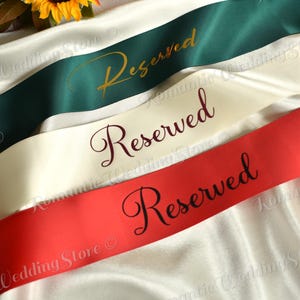 May include: Three satin ribbons in dark green, cream, and red, each featuring the word Reserved printed in a cursive script. The green ribbon has gold text, the cream ribbon has dark red text, and the red ribbon has black text.