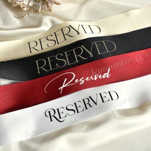 May include: Four satin sashes in cream, black, dark red, and white. The top three sashes feature the word RESERVED in a black serif font, while the third sash displays the word Reserved in a white script font. The bottom sash has RESERVED in black.