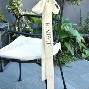 May include: A long, cream-colored satin ribbon tied in a bow at the top of a black metal chair. The ribbon features the word RESERVED printed vertically in black capital letters. The fabric has a smooth, reflective finish and hangs straight down.