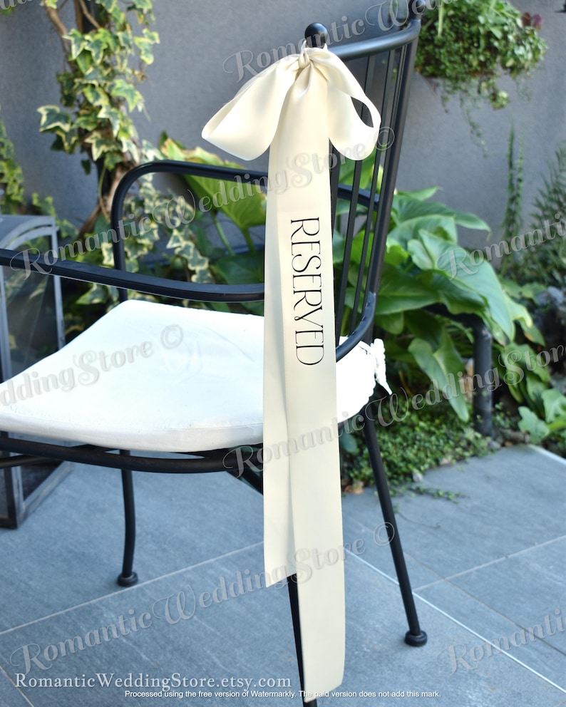 May include: A long, cream-colored satin ribbon tied in a bow at the top of a black metal chair. The ribbon features the word RESERVED printed vertically in black capital letters. The fabric has a smooth, reflective finish and hangs straight down.