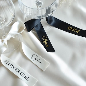 Puede incluir: Copas de champán de cristal transparente con cintas decorativas. Las cintas son blancas y negras, con letras doradas: "Flower Girl", "Sophia", "Anna" y "Bride". Sobre una superficie de satén blanco.