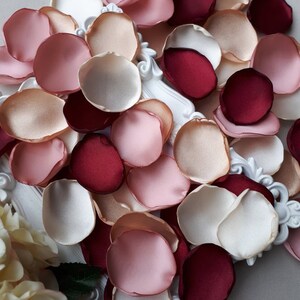 May include: Assortment of silk rose petals in ivory, blush pink, peach, and burgundy colors. The petals are arranged on a white surface.