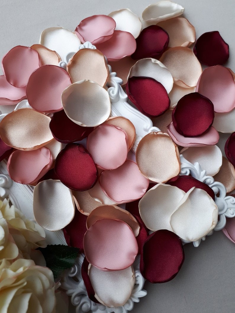 May include: Assortment of silk rose petals in ivory, blush pink, peach, and burgundy colors. The petals are arranged on a white surface.