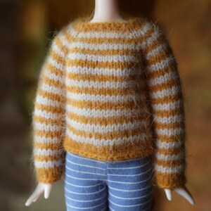 May include: A doll wearing a miniature, hand-knitted sweater with horizontal stripes in mustard yellow and white. The doll also wears blue and white striped pants. The sweater has a crew neck and long sleeves.