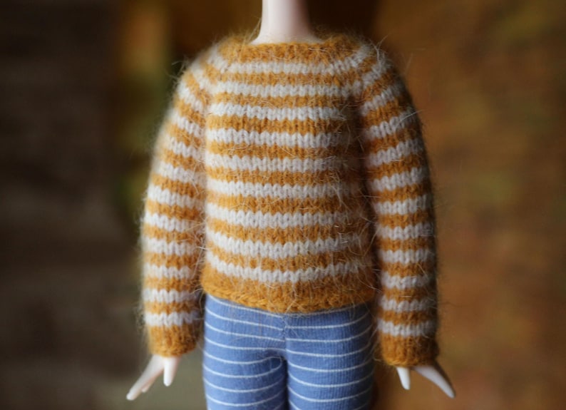 May include: A doll wearing a miniature, hand-knitted sweater with horizontal stripes in mustard yellow and white. The doll also wears blue and white striped pants. The sweater has a crew neck and long sleeves.