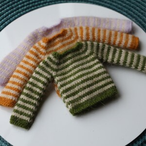 May include: Four miniature knit sweaters in various colors: lavender, orange, and green, each with horizontal stripes. The sweaters are arranged on a white surface, showcasing the handmade details and textures of the knitwear.