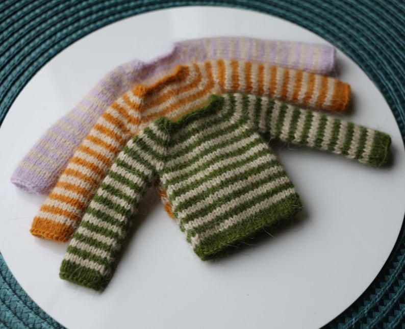 May include: Four miniature knit sweaters in various colors: lavender, orange, and green, each with horizontal stripes. The sweaters are arranged on a white surface, showcasing the handmade details and textures of the knitwear.