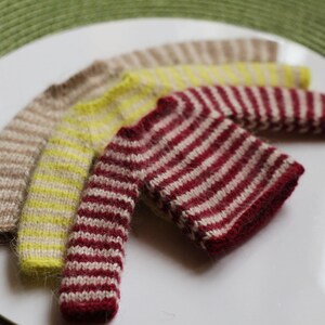 May include: Four miniature knitted sweaters in a variety of colors, including beige, yellow, and burgundy with white stripes. The sweaters are arranged on a white plate, showcasing their intricate knit patterns and textures.