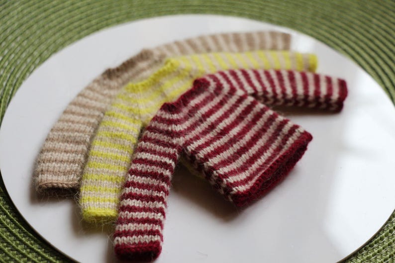May include: Four miniature knitted sweaters in a variety of colors, including beige, yellow, and burgundy with white stripes. The sweaters are arranged on a white plate, showcasing their intricate knit patterns and textures.