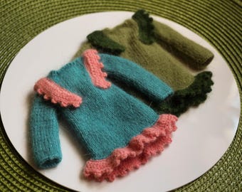 Puede incluir: Dos vestidos de punto en miniatura. Uno es turquesa con ribete rosa, el otro es verde oliva con ribete verde oscuro. Los vestidos están sobre un plato blanco, sobre un fondo verde.