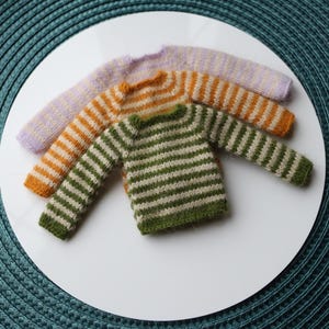 May include: Three miniature knit sweaters in a row. The sweaters are in shades of green and white, orange and white, and lavender and white stripes. The sweaters are on a white surface.