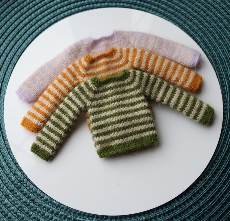 May include: Three miniature knit sweaters in a row. The sweaters are in shades of green and white, orange and white, and lavender and white stripes. The sweaters are on a white surface.