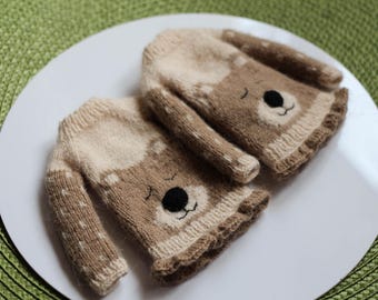 May include: Two knitted baby sweaters with a bear face design. The sweaters are primarily tan and cream-colored, with black embroidered eyes and nose. The sweaters are lying on a white plate.
