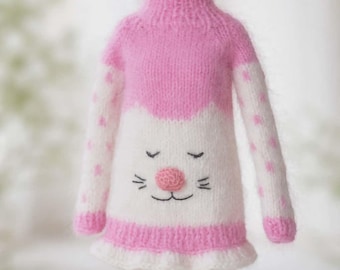 May include: A knitted sweater with a high neck, featuring a pink top section and a white bottom section. The white area displays an embroidered cat face with a raised pink nose, closed eyes, and whiskers. The sleeves have pink polka dots.