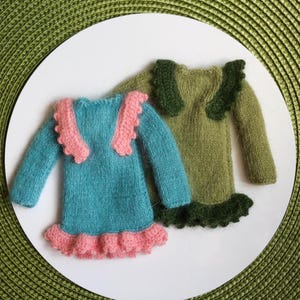 Może przedstawiać: Dwie miniaturowe dzianinowe sukienki. Jedna jest turkusowa z r&oacute;żowymi wykończeniami na kołnierzu i obszyciu. Druga jest oliwkowozielona z ciemnozielonymi wykończeniami. Obie mają długie rękawy i są prezentowane na białej powierzchni.
