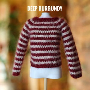 May include: A miniature burgundy and white striped sweater. The sweater is displayed on a white mannequin and has long sleeves. The text "DEEP BURGUNDY" is displayed above the sweater. The sweater is made of a fuzzy material.