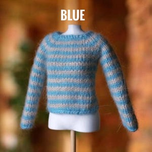 May include: A blue and light gray striped sweater for a doll. The sweater is made of a soft, fuzzy material and has long sleeves. The word "BLUE" is written in white block letters above the sweater.